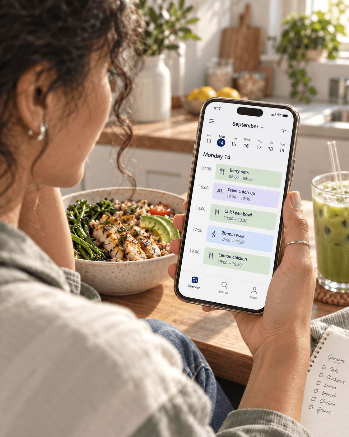 Someone checking their week in their phone calendar: berry oats, a chickpea bowl, a walk and lemon chicken, with lunch and a grocery list on the table